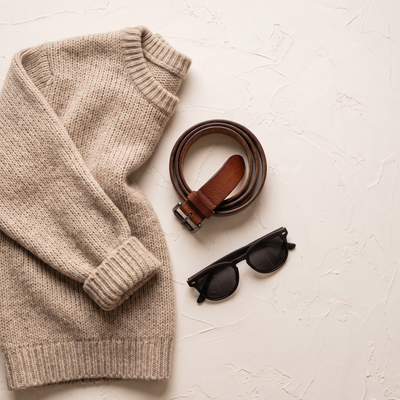 A folded knit, a leather belt and sunglasses on a pale surface